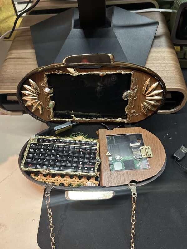 An ornate vintage handbag transformed into a tiny computer, with a black screen in its decorated lid, miniature keyboard and circuit board inside.